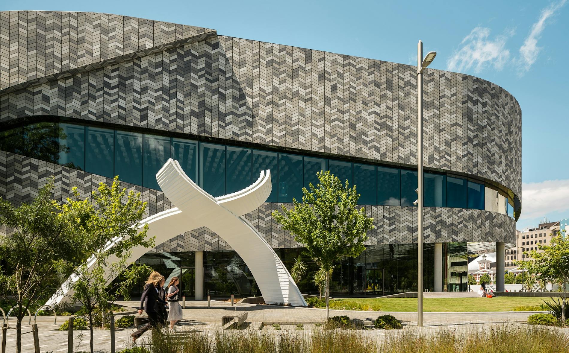 Te Pae features a distinctive architectural structure with a flowing design and a large white archway. People walk through a landscaped area under a clear blue sky, enjoying the vibrant surroundings.